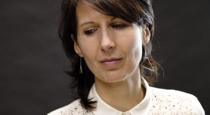 Composer Ana Sokolović is pictured against a dark background from the shoulders up. She is looking away from the camera with her head tilted to the side and is wearing a white blouse.