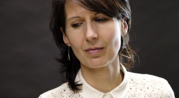 Composer Ana Sokolović is pictured against a dark background from the shoulders up. She is looking away from the camera with her head tilted to the side and is wearing a white blouse.