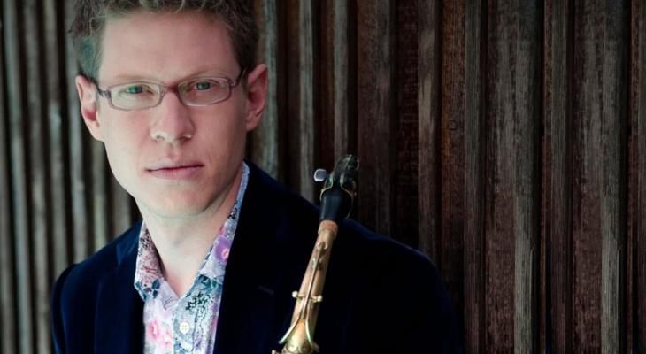 Wallace Halladay leans against a wooden background wall. He is wearing a black suit jacket and a floral shirt, holding a saxophone in his hand.