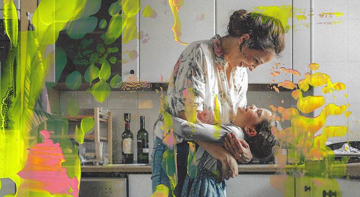 A photograph of a mother and daughter hugging in a kitchen with splatters and smears of yellow and pink paint.