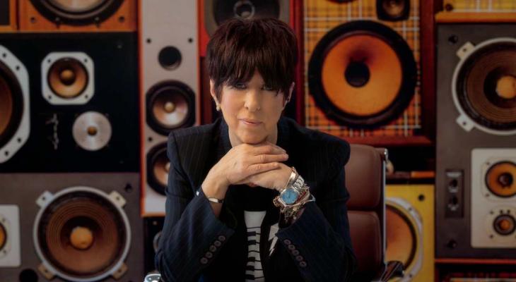 A portrait of Diane Warren seated in front of a wall of speakers.