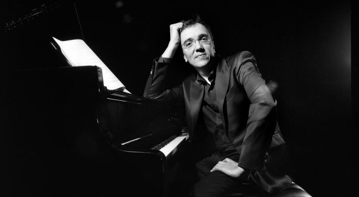 Éric Le Sage is sitting at the piano, leaning on the instrument and looking at the camera, smiling.
