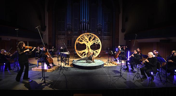 An actor poses in front of a wooden carving of a maple tree, centre stage under a bright light, with Tafelmusik musicians performing around them.