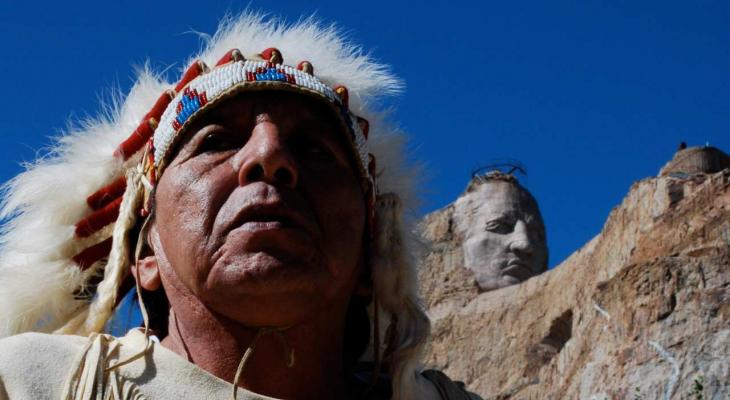 An Indigenous man in a headdress appears in front of Mount Rushmore.