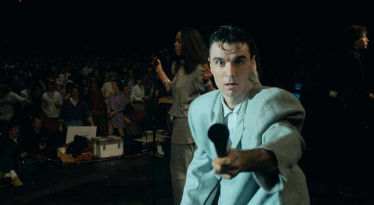 David Byrne reaches out a microphone towards the camera.