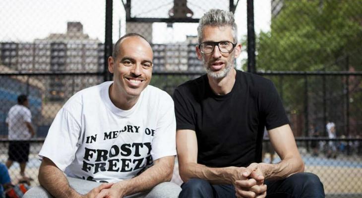 DJs Stretch and Bobbito smile for the camera outside a basketball court. 