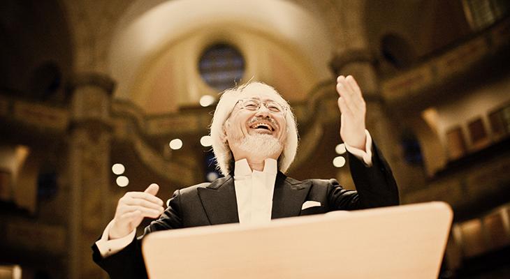 Close-up of Masaaki Suzuki conducting from the podium. He is an older Asian male, smiling and wearing a black tuxedo and white white with a high collar.