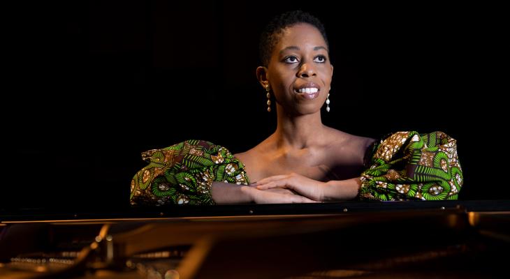 Dr. Ege is seated, leaning on a grand piano, smiling and looking into the distance. She is a Black woman wearing a green, yellow and black patterned top and dangling earrings.