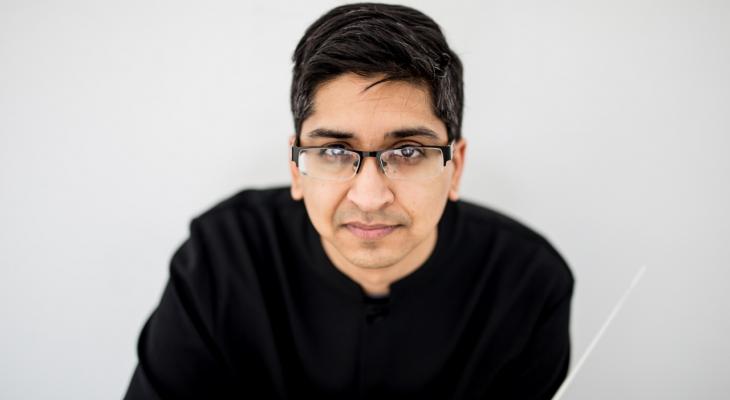 Pratik Gandhi is in front of a white background. He is wearing glasses and a black sweater.