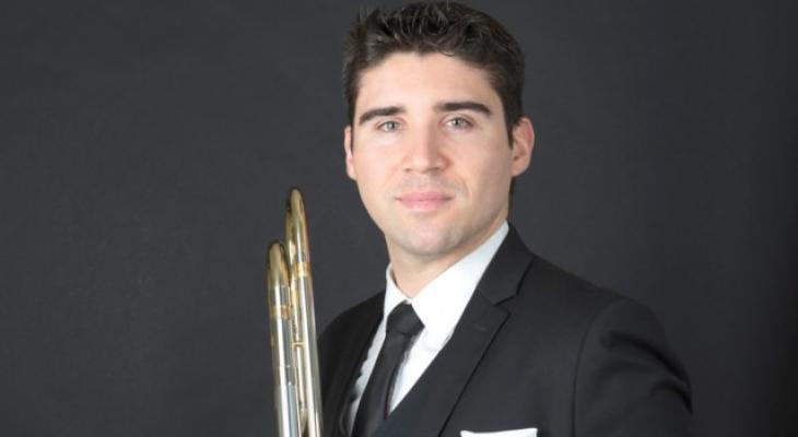 Trombonist Fabrice Millischer, against a black background, dressed in a black suit, wearing a tie, holding a trombone.