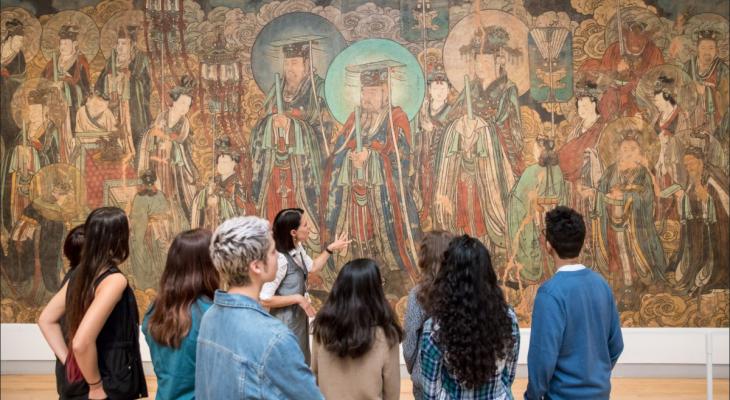 Tour Guide speaks to a group of 8 people standing in front of a large Chinese mural.