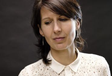Composer Ana Sokolović is pictured against a dark background from the shoulders up. She is looking away from the camera with her head tilted to the side and is wearing a white blouse.