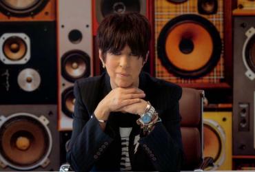 A portrait of Diane Warren seated in front of a wall of speakers.