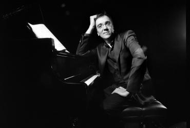 Éric Le Sage is sitting at the piano, leaning on the instrument and smiling at the camera.