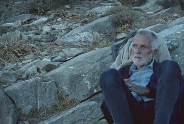 a man with a beard sitting next to a large rock