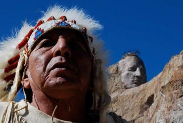 An Indigenous man in a headdress appears in front of Mount Rushmore.