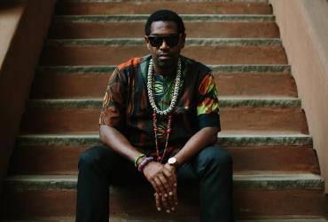 Yosvany Terry is seated on steps looking straight at the camera. He is wearing sunglasses, a multicoloured shirt and dark pants, and beaded jewellery around his neck and wrist.