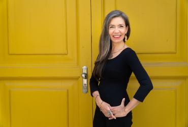 Patricia Caicedo standing in front of a yellow door, smiling, wearing a black dress, with her hair down.