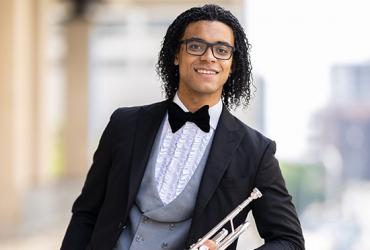 William Leathers outdoors, dressed in a black suit, wearing a bowtie, glasses, holding a trumpet, smiling at the camera.