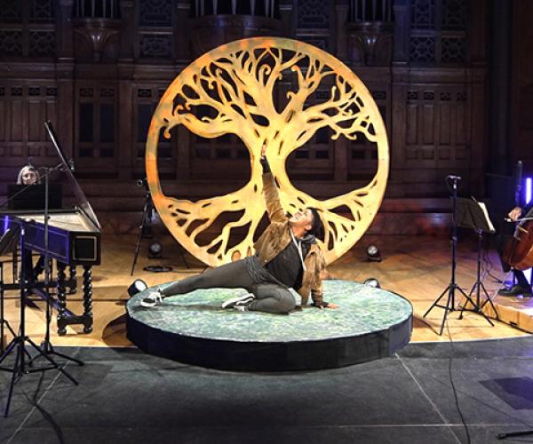 An actor poses in front of a wooden carving of a maple tree, centre stage under a bright light, with Tafelmusik musicians performing around them.